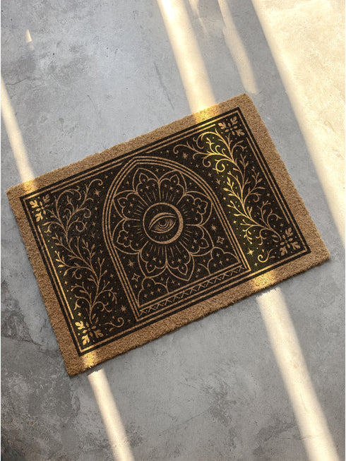 A rectangular, natural brown coir doormat with a non-slip black vinyl backing, viewed from directly above. The mat is made of textured coconut fiber. The design features intricate black line art in a Victorian Gothic and Art Nouveau style. In the center is an All Seeing Eye, or Eye of Providence, enclosed within a radiant starburst and a mandala-like floral pattern. This central motif is set inside an arched frame. The corners and sides are adorned with symmetrical, flowing botanical vines.